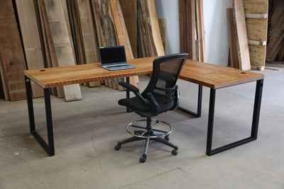 Modern L-shaped wooden desk with metal legs, paired with a black mesh ergonomic chair and a laptop
